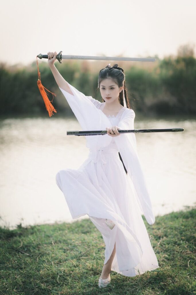 Asian woman practicing Tai Chi sword kata in a white dress at a serene park setting.